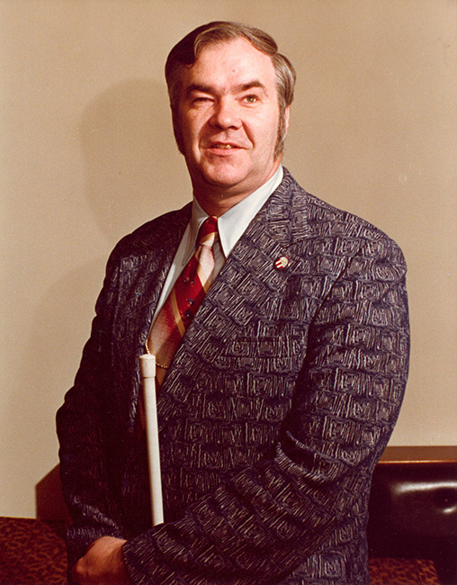 Oral Miller, pictured standing, in a dark suit coat, white shirt, and red- and gold-striped tie, smiles for the camera. Photo taken circa 1980.